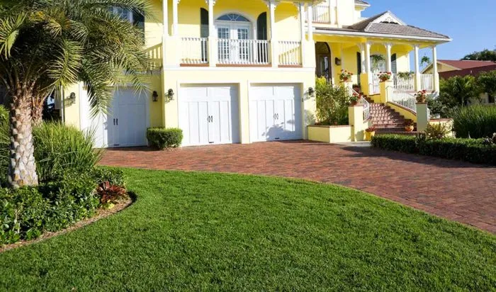 A yellow two-story house with a white double garage, balcony, and a brick driveway is surrounded by a well-maintained lawn courtesy of Sanford Lawn Care and lush tropical vegetation.