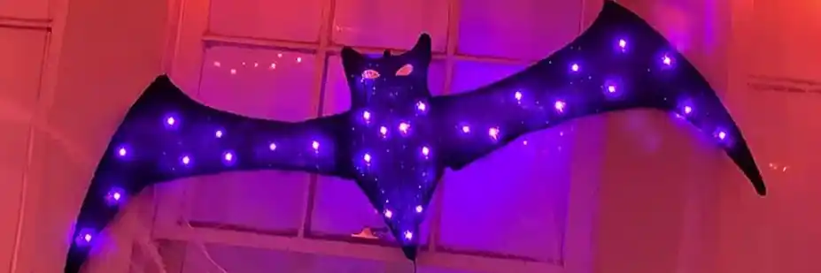 A large purple bat decoration with glowing lights across its wings, hanging in front of a window, representing Halloween decor and awareness for bat exclusion practices.